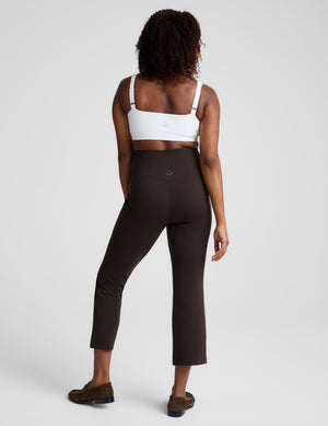 A woman shown from the back wearing high-waisted dark brown cropped leggings and a white sports bra, standing against a light gray studio background.
