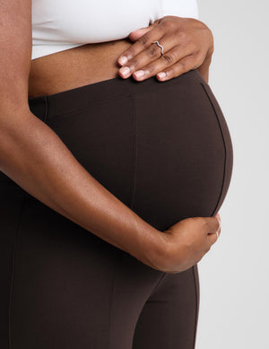 Close-up of a pregnant person wearing dark brown maternity leggings, holding their belly. The person is also wearing a white bra, and the background is a clean, light gray studio backdrop.