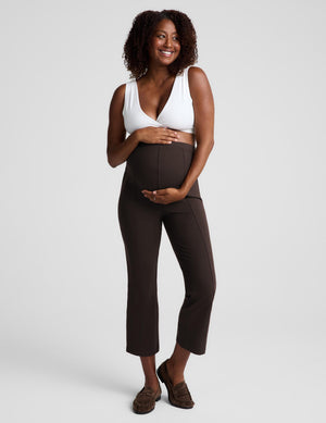 A smiling pregnant woman stands against a light gray studio background wearing dark brown maternity pants and a white bra, with her hands resting on her belly.