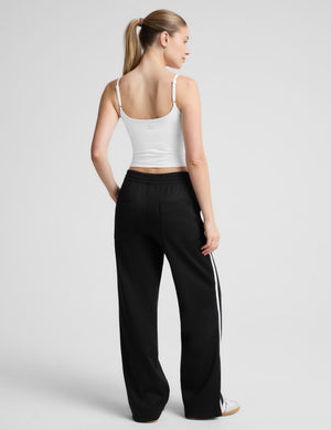 A woman modeling a white spaghetti-strap tank top and black wide-leg track pants against a light gray studio background.