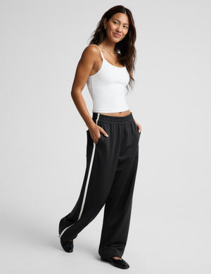 A woman modeling black wide-leg sweatpants with a white side stripe, paired with a white ribbed camisole top, on a light gray studio background.