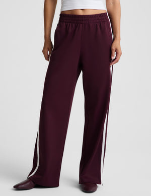 A model wearing burgundy wide-leg sweatpants with white side stripes, paired with a white crop top and dark brown pointed-toe shoes, photographed against a light gray studio background.