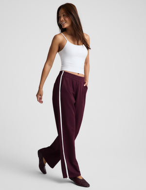 A woman modeling a pair of wide-leg burgundy sweatpants with a white side stripe, paired with a white ribbed tank top and dark flats, against a light gray studio background.