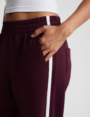 Close-up of a person wearing burgundy athletic pants with a white side stripe and white waistband detail, shown against a light gray background.