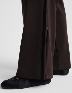 Close-up of dark brown wide-leg trousers with a black side zipper detail, shown on a model wearing black flats against a light gray studio background.