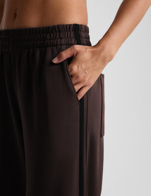 Woman wearing brown track pants with black flat shoes against a light gray background. 
