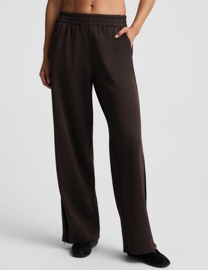 Woman wearing brown track pants with black flat shoes against a light gray background. 