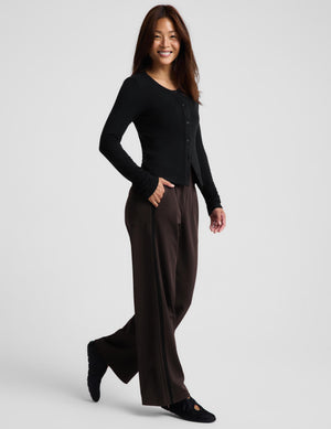 Woman wearing a black cardigan and brown track pants with black flat shoes against a light gray background. 