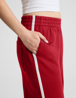 A close-up of a person wearing red track pants with a white side stripe, showing the side pocket and hand in the pocket, paired with a white cropped top against a light gray background.