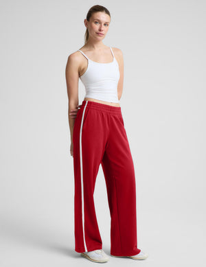A woman modeling red wide-leg athletic pants with a white side stripe, paired with a white tank top and white sneakers, against a light gray studio background.