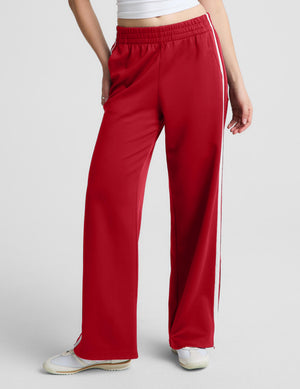 A person wearing red wide-leg athletic pants with white side stripes, paired with a white cropped top and white sneakers, photographed against a light gray studio background.