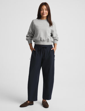 A woman modeling a light gray crewneck sweatshirt and dark navy wide-leg drawstring pants, paired with brown loafers, against a solid light gray studio background.