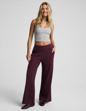 A woman modeling a pair of wide-leg, high-waisted pants in a deep burgundy, paired with a light gray ribbed crop top and dark flats, against a solid light gray studio background.