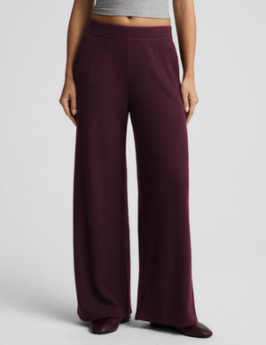 A model wearing a pair of wide-leg knit pants in a deep burgundy, paired with a gray crop top and dark brown shoes, against a light gray studio background.