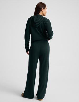A woman modeling a dark forest green zip-up hoodie and matching wide-leg sweatpants, styled with a white sports bra and tan slip-on shoes against a light gray studio background.