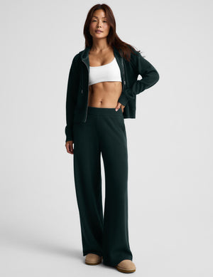 A woman modeling a dark forest green zip-up hoodie and matching wide-leg sweatpants, styled with a white sports bra and tan slip-on shoes against a light gray studio background.