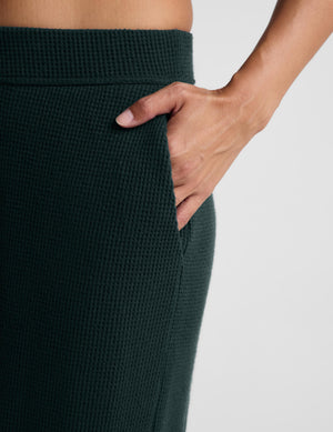 Close-up of a person wearing dark forest green waffle-knit pants, showing a hand tucked into a side pocket against a light gray background.