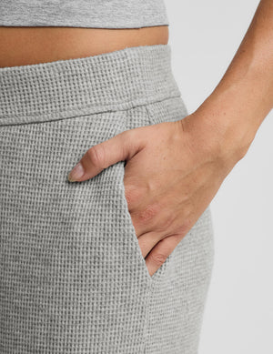 Close-up of a person wearing light gray waffle-knit lounge pants, showing a hand tucked into the side pocket.