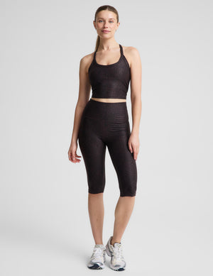 A woman modeling a matching activewear set consisting of a dark brown leopard crop tank and high-waisted pedal pushers, paired with white athletic sneakers, against a light gray studio background.