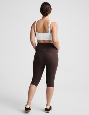 A woman shown from the back wearing dark brown high-waisted capri leggings and a white sports bra, standing against a light gray studio background.