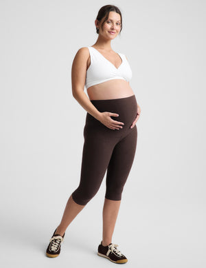 A pregnant woman posing in a white sports bra and dark brown maternity capri leggings, paired with black and white sneakers, against a light gray studio background.