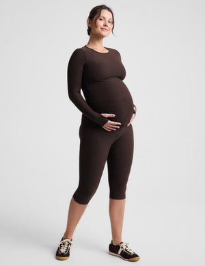 A pregnant woman modeling a matching dark brown long-sleeve top and capri leggings set, standing against a light gray studio background.