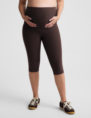A model wearing dark brown maternity capri leggings and brown sneakers, standing against a light gray studio background.