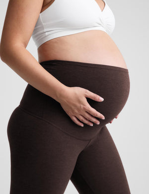 Pregnant woman wearing a white sports bra and dark brown maternity leggings, holding her baby bump against a light gray background.