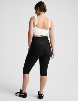 Pregnant woman wearing a white maternity bra and black pedal pushers against a plain background. 