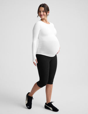 Pregnant woman wearing a white long sleeve maternity top and black pedal pushers against a plain background. 
