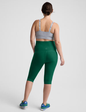 A woman is shown from the back wearing dark green high-waisted capri leggings, a gray sports bra, and blue athletic sneakers against a light gray studio background.