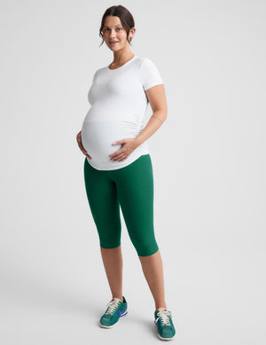 A pregnant woman modeling a white short-sleeve crewneck top and green capri leggings with teal sneakers, standing against a light gray studio background.