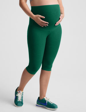 A model wearing high-waisted green maternity capri leggings and green sneakers, standing against a light gray studio background.