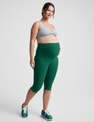 A pregnant model wearing high-waisted green maternity capri leggings, a light gray sports bra, and green sneakers, standing against a light gray studio background.