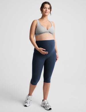 A pregnant model wearing a light gray crossover sports bra and navy blue maternity capri leggings, standing against a light gray studio background.