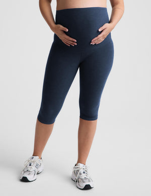 A pregnant model wearing a light gray crossover sports bra and navy blue maternity capri leggings, standing against a light gray studio background.