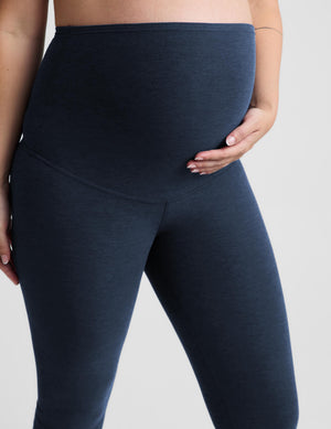 A pregnant model wearing a light gray crossover sports bra and navy blue maternity capri leggings, standing against a light gray studio background.
