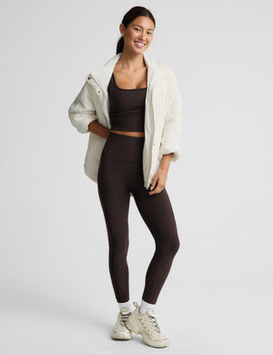 A woman modeling a dark brown activewear set consisting of a sports bra and high-waisted leggings, styled with an open cream-colored lightweight jacket and beige sneakers against a light gray studio background.