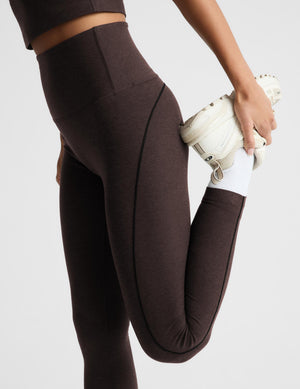 A close-up of a person wearing dark brown high-waisted leggings and a matching sports bra, stretching their leg while holding a white sneaker against a light gray studio background.