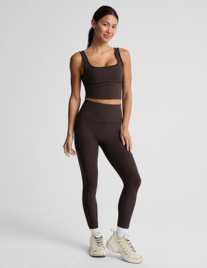A woman modeling a matching dark brown activewear set consisting of a cropped tank top and high-waisted leggings, paired with white socks and beige sneakers, against a light gray studio background.