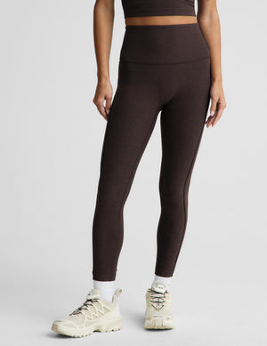 A model wearing dark brown high-waisted leggings and cream-colored athletic sneakers, shown from the waist down against a light gray studio background.