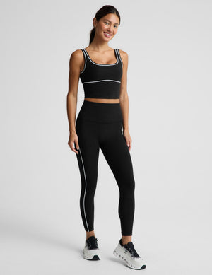 A woman modeling a black athletic set consisting of a cropped tank top and high-waisted leggings with white contrast piping details, paired with white and black sneakers, against a light gray studio background.