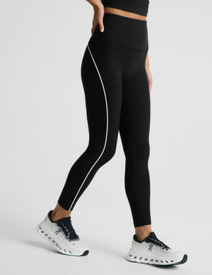 Black high-waisted athletic leggings with a white side stripe, modeled on a person wearing white and black running shoes against a light gray studio background.