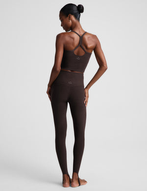 A woman modeling a matching dark brown activewear set consisting of a cropped tank and high-waisted stirrup leggings, posed against a light gray studio background.
