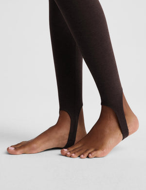 A woman modeling a matching dark brown activewear set consisting of a cropped tank and high-waisted stirrup leggings, posed against a light gray studio background.