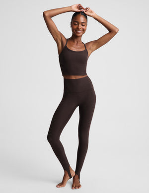 A woman modeling a matching dark brown activewear set consisting of a cropped tank and high-waisted stirrup leggings, posed against a light gray studio background.