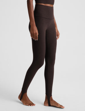 A woman modeling a matching dark brown activewear set consisting of a cropped tank and high-waisted stirrup leggings, posed against a light gray studio background.