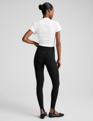 A woman modeling black leggings, a white top, and a black draped jacket, standing against a light gray studio background.