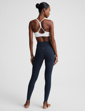 A woman shown from the back wearing high-waisted navy blue leggings and a white sports bra, standing against a light gray studio background.