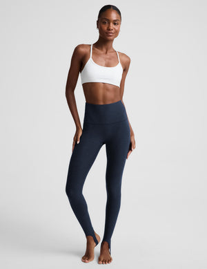 A woman modeling high-waisted navy blue leggings and a white sports bra against a light gray studio background.